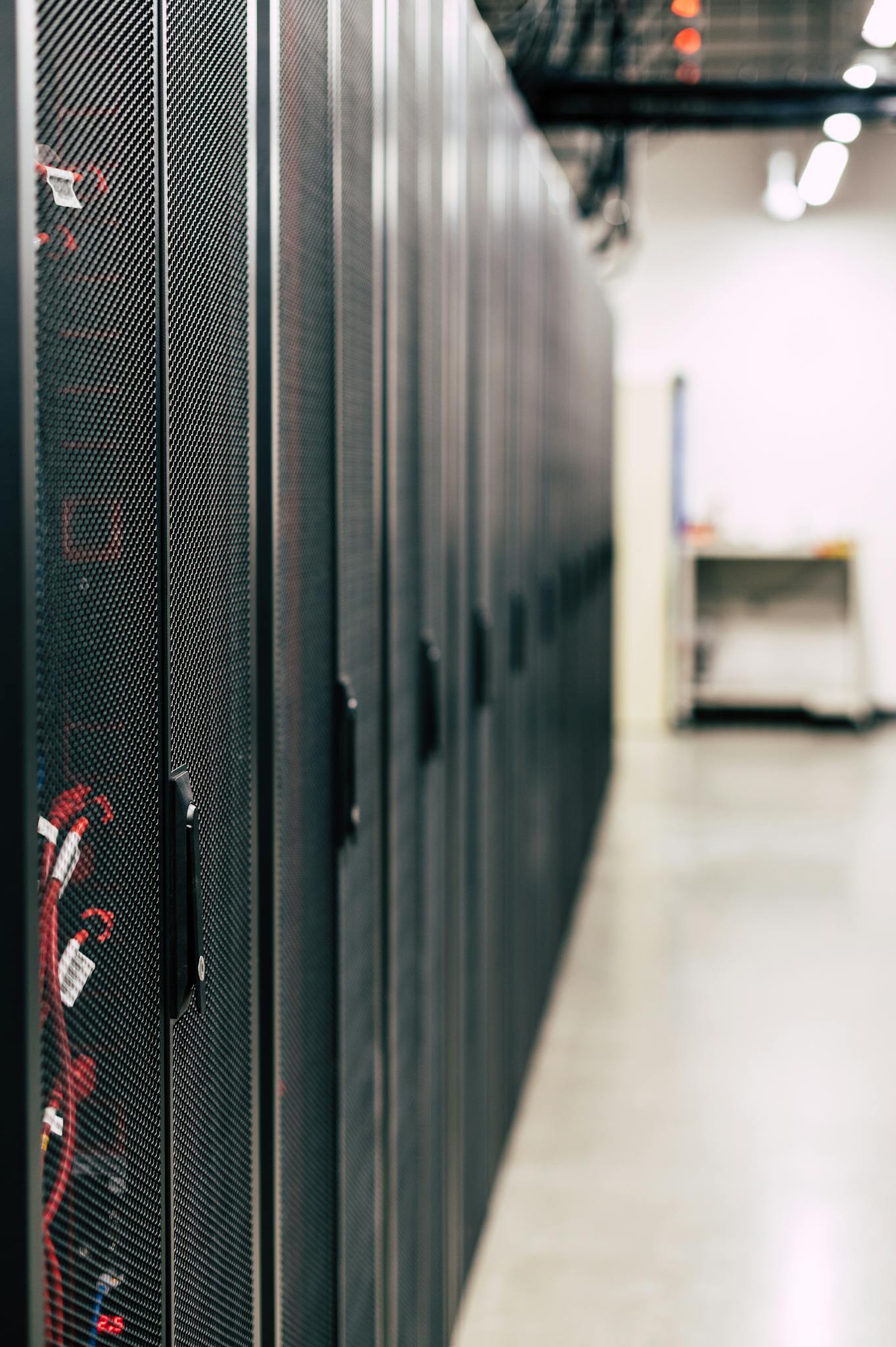 Metal black cabinets with servers inside with red wires connected standing in row in lighted room